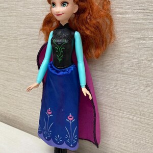 May include: A doll of a character with red hair, wearing a black bodice with green accents, a blue skirt with floral designs, and a purple cape. The doll has a smiling expression and is standing.