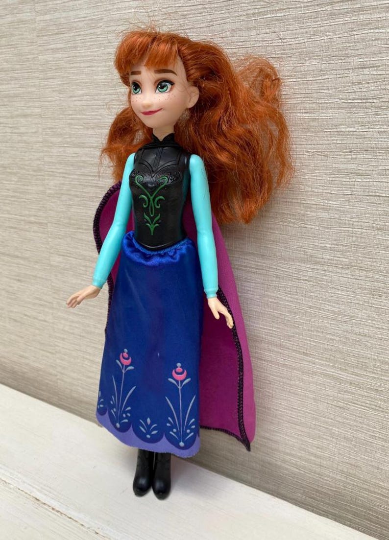 May include: A doll of a character with red hair, wearing a black bodice with green accents, a blue skirt with floral designs, and a purple cape. The doll has a smiling expression and is standing.