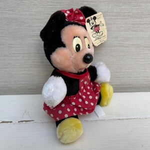 K&ouml;nnte beinhalten: Eine Minnie Mouse Pl&uuml;schpuppe mit schwarzem Fell, einem roten Kleid mit wei&szlig;en Punkten und einer passenden Schleife. Die Puppe hat wei&szlig;e Handschuhe und gelbe Schuhe. Ein Etikett mit einer Disney-Charakter-Illustration ist angebracht.