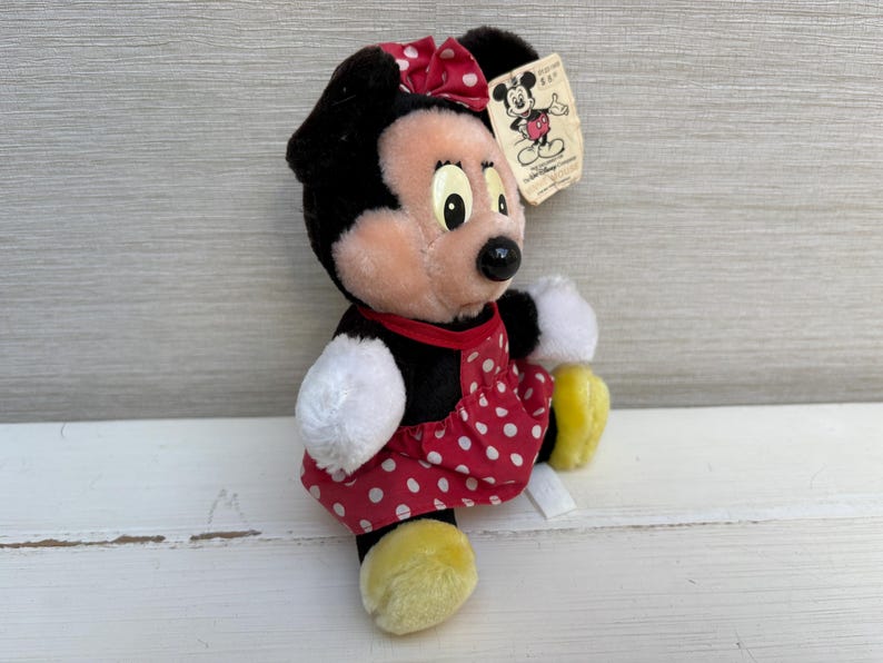 K&ouml;nnte beinhalten: Eine Minnie Mouse Pl&uuml;schpuppe mit schwarzem Fell, einem roten Kleid mit wei&szlig;en Punkten und einer passenden Schleife. Die Puppe hat wei&szlig;e Handschuhe und gelbe Schuhe. Ein Etikett mit einer Disney-Charakter-Illustration ist angebracht.