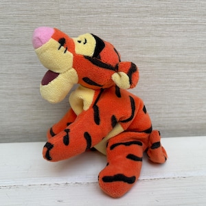 May include: A plush toy tiger in a sitting position. The tiger is orange with black stripes, a pink nose, and yellow accents. The toy has a friendly expression and is made of soft fabric.