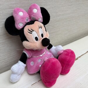 May include: A plush Minnie Mouse doll, featuring a pink dress with white polka dots, matching bow with white polka dots, and large pink feet. The doll has black ears, a black nose, and white gloves. The doll is sitting on a white surface.