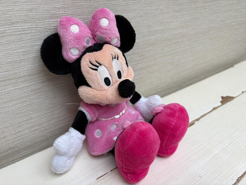 May include: A plush Minnie Mouse doll, featuring a pink dress with white polka dots, matching bow with white polka dots, and large pink feet. The doll has black ears, a black nose, and white gloves. The doll is sitting on a white surface.