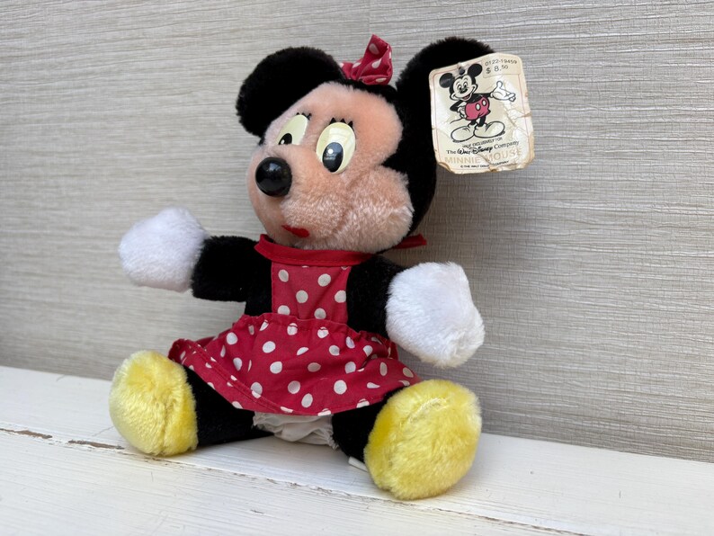 K&ouml;nnte beinhalten: Ein Vintage-Minnie-Mouse-Pl&uuml;schtier. Das Pl&uuml;schtier hat ein rot-wei&szlig; gepunktetes Kleid, schwarze Ohren und eine rote Schleife. Das Spielzeug hat ein Etikett mit einer Disney-Charakterillustration.