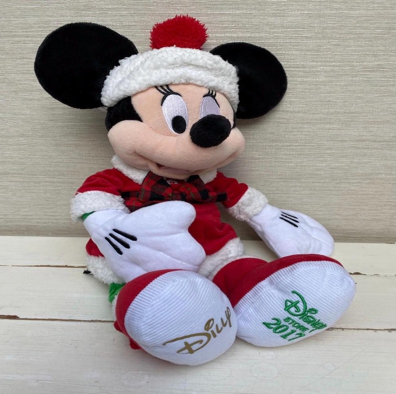 May include: A plush Minnie Mouse doll wearing a red and white Santa-style outfit with a matching hat and a red pom-pom. The doll has black ears, white gloves, and red and white striped feet with the Disney Store 2017 logo.