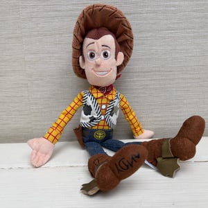 May include: A plush toy of a cowboy character with a brown hat, yellow plaid shirt, and blue jeans. The toy has a white vest with a black and white cow pattern, a red neckerchief, and brown boots. The word "ANDY" is printed on one boot.