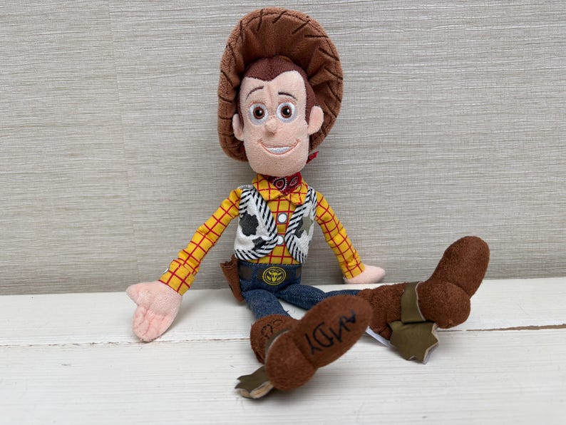 May include: A plush toy of a cowboy character with a brown hat, yellow plaid shirt, and blue jeans. The toy has a white vest with a black and white cow pattern, a red neckerchief, and brown boots. The word "ANDY" is printed on one boot.