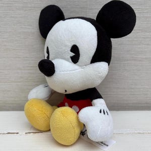 Op de afbeelding: Een pluche Mickey Mouse-speelgoed. Het iconische personage heeft een wit gezicht, zwarte oren en een zwarte neus. Hij draagt een rood shirt met witte knopen, gele schoenen en witte handschoenen. Het speelgoed staat op een wit oppervlak.