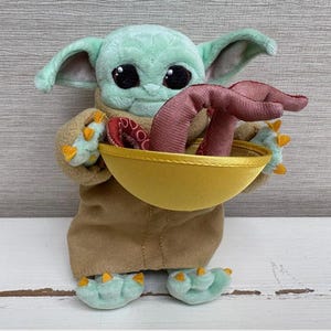 Pode incluir: Uma boneca de pel&uacute;cia de um personagem de uma popular s&eacute;rie de fic&ccedil;&atilde;o cient&iacute;fica. A boneca tem olhos grandes e pretos, orelhas grandes e segura uma tigela amarela com um objeto vermelho dentro. A boneca est&aacute; vestindo um casaco marrom e tem p&eacute;s verdes com garras amarelas.