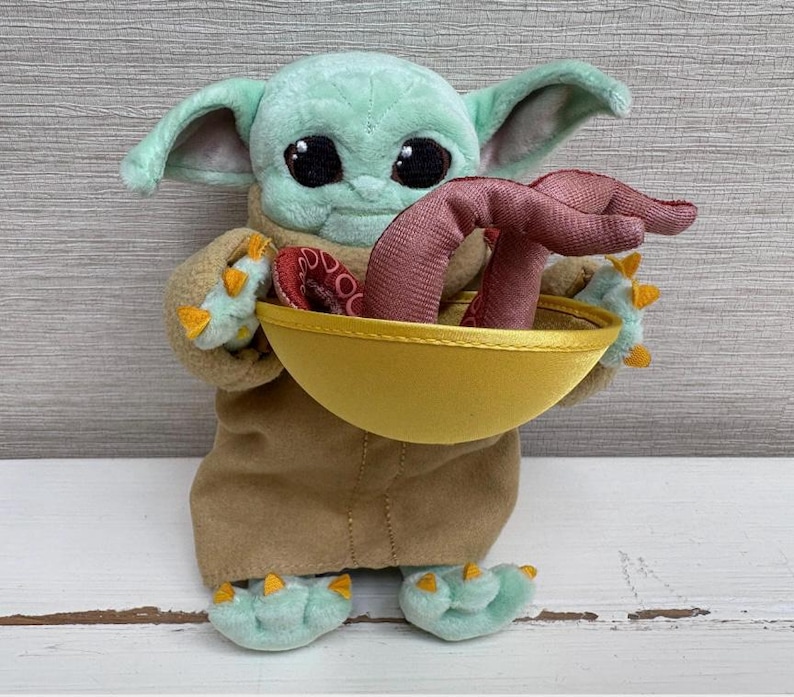 Pode incluir: Uma boneca de pel&uacute;cia de um personagem de uma popular s&eacute;rie de fic&ccedil;&atilde;o cient&iacute;fica. A boneca tem olhos grandes e pretos, orelhas grandes e segura uma tigela amarela com um objeto vermelho dentro. A boneca est&aacute; vestindo um casaco marrom e tem p&eacute;s verdes com garras amarelas.
