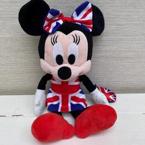 Puede incluir: Un peluche de Minnie Mouse con un lazo y un vestido con la bandera brit&aacute;nica. La mu&ntilde;eca tiene orejas negras, cara rosa, guantes blancos y zapatos rojos. El dise&ntilde;o es una representaci&oacute;n patri&oacute;tica del Reino Unido.