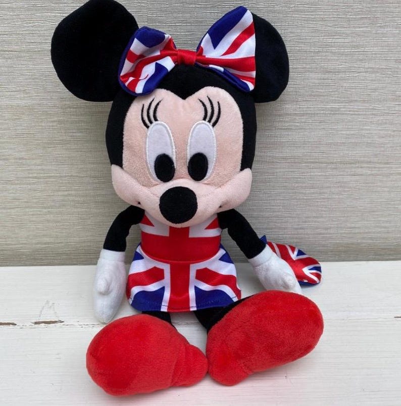 Puede incluir: Un peluche de Minnie Mouse con un lazo y un vestido con la bandera brit&aacute;nica. La mu&ntilde;eca tiene orejas negras, cara rosa, guantes blancos y zapatos rojos. El dise&ntilde;o es una representaci&oacute;n patri&oacute;tica del Reino Unido.