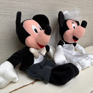 May include: Two plush Disney characters, Mickey and Minnie Mouse, dressed for a wedding. Mickey wears a black tuxedo with a white bow tie. Minnie is in a white wedding dress and veil. Both have black ears and red mouths.