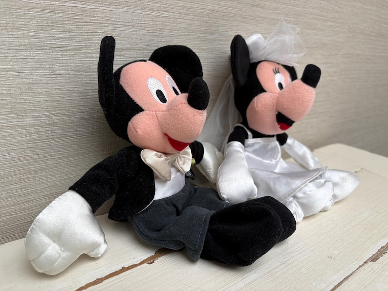 May include: Two plush Disney characters, Mickey and Minnie Mouse, dressed for a wedding. Mickey wears a black tuxedo with a white bow tie. Minnie is in a white wedding dress and veil. Both have black ears and red mouths.