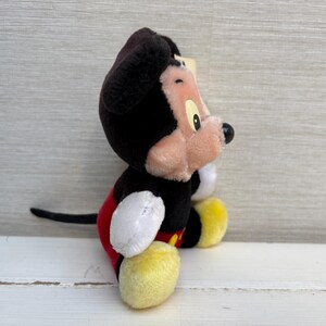 Puede incluir: Un peluche de Mickey Mouse, con cabeza y orejas negras, cara rosa y nariz negra. El juguete tiene guantes blancos, zapatos amarillos y pantalones cortos rojos. El personaje cl&aacute;sico de Disney est&aacute; sentado.