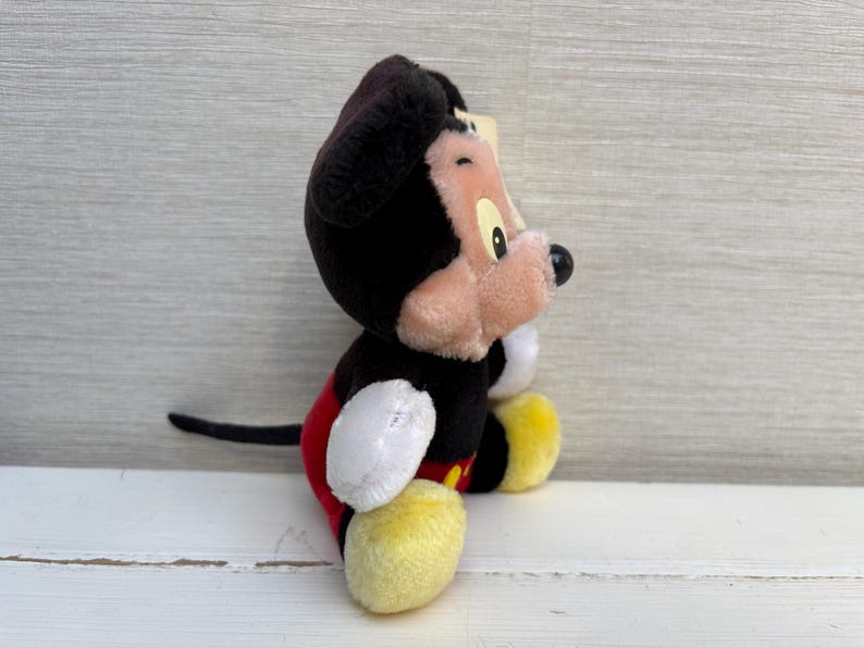 Puede incluir: Un peluche de Mickey Mouse, con cabeza y orejas negras, cara rosa y nariz negra. El juguete tiene guantes blancos, zapatos amarillos y pantalones cortos rojos. El personaje cl&aacute;sico de Disney est&aacute; sentado.