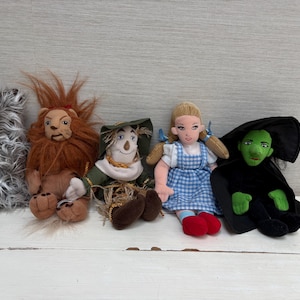 Könnte beinhalten: Ein Set aus fünf Plüschpuppen, bestehend aus einem grauen zotteligen Hund, einem braunen Löwen mit Mähne, einer Vogelscheuche mit grünem Hut, einer blonden Puppe in einem blau-weiß karierten Kleid mit roten Schuhen und einer grünen Figur im schwarzen Umhang.