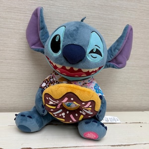 Op de afbeelding: Een blauwe Stitch-knuffel met grote oren en een ondeugende uitdrukking, die een donut vasthoudt met chocoladeglazuur en hagelslag. De pop draagt een shirt met een patroon. Het pluche speelgoed zit op een wit oppervlak.