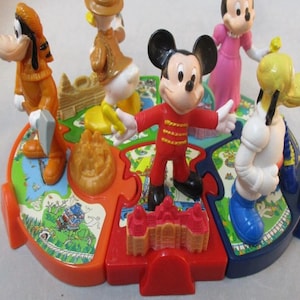 Könnte beinhalten: Ein farbenfrohes Disney-Charakterspielzeug mit Mickey Mouse, Minnie Mouse, Goofy, Donald Duck und Pluto Figuren. Das Spielzeug hat eine kreisförmige Basis mit Puzzleteilen, jeweils mit einer thematischen Illustration. Die Charaktere sind in verschiedenen Posen.