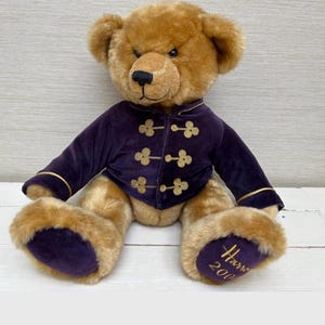 May include: A teddy bear with tan fur, dressed in a purple velvet jacket with gold buttons and trim. The bear has a black nose and is seated. The bear's foot has the text "Harrods 2000".