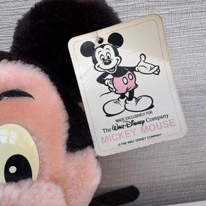 Puede incluir: Primer plano de un peluche vintage de Mickey Mouse con una etiqueta. El peluche tiene una cara rosa, ojos blancos grandes y orejas negras. La etiqueta presenta una ilustraci&oacute;n de dibujos animados de Mickey Mouse y dice "Made exclusively for The Walt Disney Company MICKEY MOUSE".