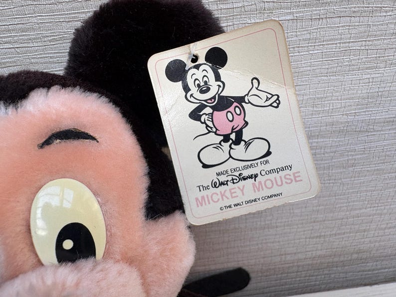 Puede incluir: Primer plano de un peluche vintage de Mickey Mouse con una etiqueta. El peluche tiene una cara rosa, ojos blancos grandes y orejas negras. La etiqueta presenta una ilustraci&oacute;n de dibujos animados de Mickey Mouse y dice "Made exclusively for The Walt Disney Company MICKEY MOUSE".