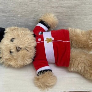 May include: A plush toy bear wearing a red jacket with a white stripe and gold emblem, black hat, and tan fur. The bear is lying down on a white wooden surface.