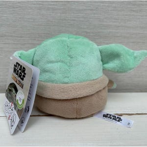 May include: A Grogu plush toy, also known as Baby Yoda, from The Mandalorian. The toy is light green with large ears, a tan outfit, and a brown base. A Star Wars tag is attached.