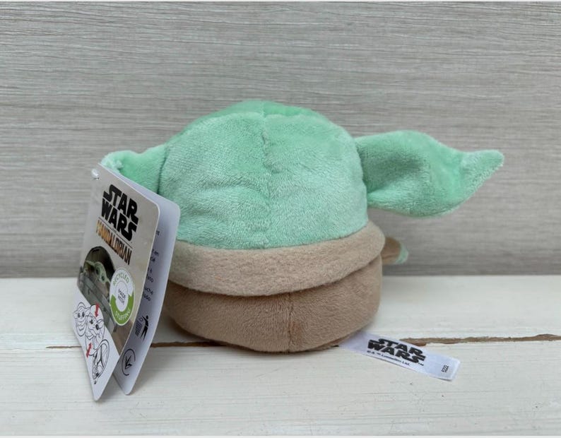 May include: A Grogu plush toy, also known as Baby Yoda, from The Mandalorian. The toy is light green with large ears, a tan outfit, and a brown base. A Star Wars tag is attached.