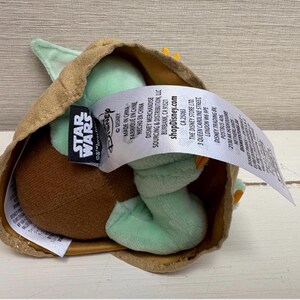 May include: A Grogu plush toy, also known as Baby Yoda, inside a tan-coloured carrier. The doll features large green ears, a brown head, and a light green body. A Star Wars tag is visible.
