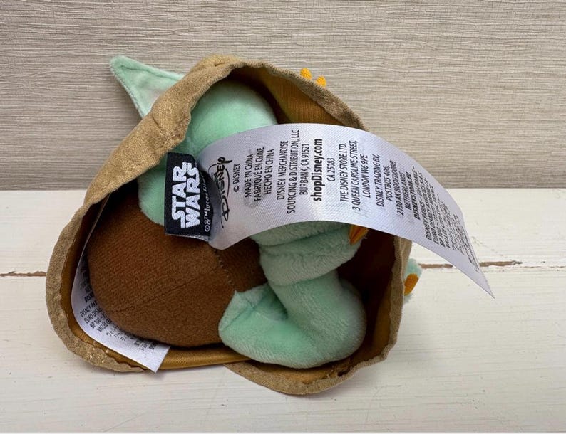 May include: A Grogu plush toy, also known as Baby Yoda, inside a tan-coloured carrier. The doll features large green ears, a brown head, and a light green body. A Star Wars tag is visible.