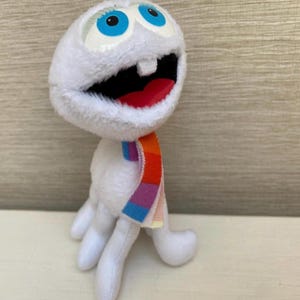 Peut inclure: Une peluche blanche avec de grands yeux bleus, une large bouche ouverte avec une langue rouge et une écharpe colorée. Le jouet a un design fantaisiste et une expression amicale, idéal pour les enfants.