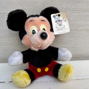 Puede incluir: Un peluche de Mickey Mouse con orejas negras, cuerpo negro, pantalones cortos rojos y zapatos amarillos. El juguete tiene una etiqueta con una ilustraci&oacute;n de Mickey Mouse y el texto "The Walt Disney Company". El juguete est&aacute; sobre una superficie blanca.