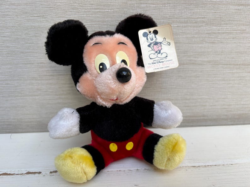 Puede incluir: Un peluche de Mickey Mouse con orejas negras, cuerpo negro, pantalones cortos rojos y zapatos amarillos. El juguete tiene una etiqueta con una ilustraci&oacute;n de Mickey Mouse y el texto "The Walt Disney Company". El juguete est&aacute; sobre una superficie blanca.
