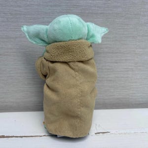 May include: A plush toy of a character with large ears and a tan coat. The toy has a light green head and ears, with a tan coat and a brown collar. The toy is made of soft fabric and is designed to resemble a popular character.