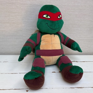 May include: A plush toy of a green turtle character wearing a red eye mask, a tan chest plate, and dark red fabric accents on the arms, legs, and waist. The character has embroidered green eyes and a stitched smile, with a soft, stuffed body.