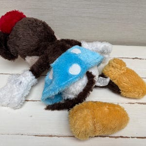 May include: A plush toy in the shape of a cartoon character, featuring a brown body, red pom-pom, blue and white accents, and tan feet. The toy is lying on a light-colored wooden surface.