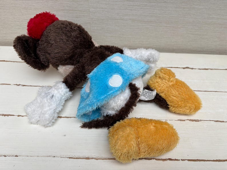 May include: A plush toy in the shape of a cartoon character, featuring a brown body, red pom-pom, blue and white accents, and tan feet. The toy is lying on a light-colored wooden surface.