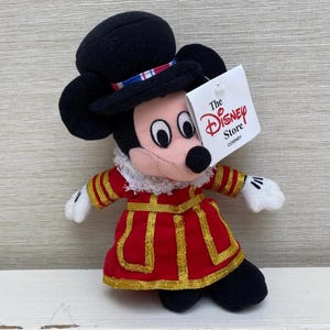 May include: A Mickey Mouse soft toy dressed in a red and gold uniform and a black hat. The toy has a tag that reads "The Disney Store". A collectible item.