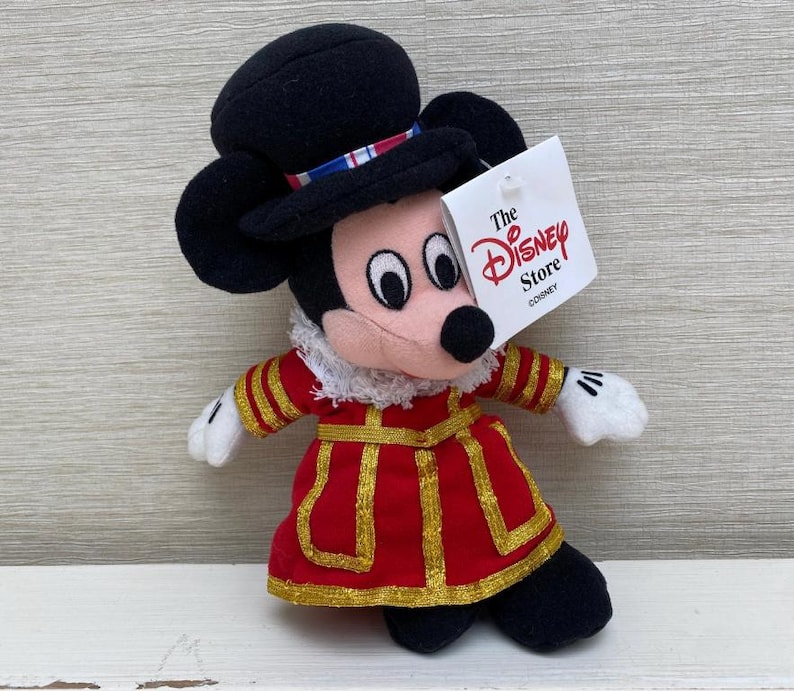 May include: A Mickey Mouse soft toy dressed in a red and gold uniform and a black hat. The toy has a tag that reads "The Disney Store". A collectible item.