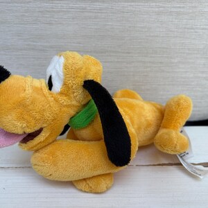 Op de afbeelding: Een knuffel van het personage Pluto, met goudgele vacht, lange zwarte oren, een zwarte neus en een felgroene halsband. Het knuffeldier ligt in een liggende positie met de roze tong naar buiten en een wit label aan de zijkant bevestigd.