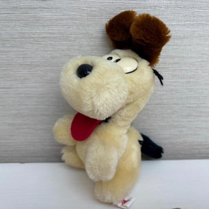 May include: A soft toy dog with a cream-coloured body, brown ears, and a black tail. The dog has a red tongue and black eyes. The toy is standing on a white surface against a grey background.