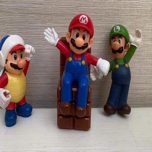 May include: Three colorful Mario Brothers figurines. Mario is seated on a brown brick block, wearing a red hat and blue overalls. Luigi is standing, wearing a green hat and overalls. A third character is also present.
