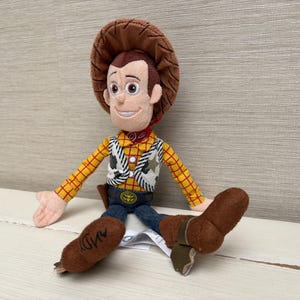 May include: A plush Woody doll from the Toy Story franchise. The doll features a brown cowboy hat, a yellow plaid shirt, a white vest with black cow print, blue jeans, and brown boots. The doll has a red neckerchief and a sheriff's badge.