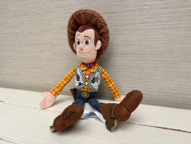 May include: A plush Woody doll from the Toy Story franchise. The doll features a brown cowboy hat, a yellow plaid shirt, a white vest with black cow print, blue jeans, and brown boots. The doll has a red neckerchief and a sheriff's badge.