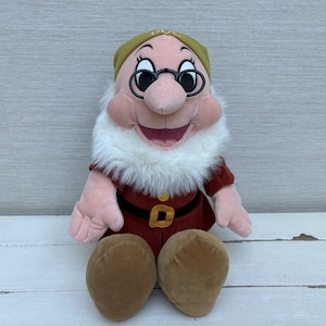 Op de afbeelding: Een pluche pop van het personage Doc uit Sneeuwwitje en de zeven dwergen. De pop heeft een witte baard, een bril en een rood en bruin kostuum. De pop zit op een wit oppervlak.