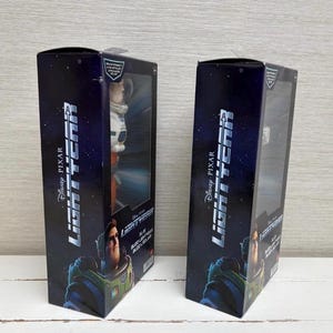 May include: Two black boxes with the Disney Pixar Lightyear logo and character images. Each box has a clear plastic window displaying a Buzz Lightyear action figure. The boxes are on a white wooden surface.