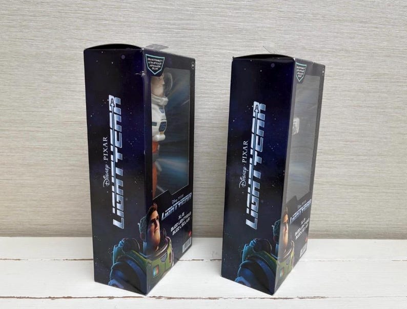 May include: Two black boxes with the Disney Pixar Lightyear logo and character images. Each box has a clear plastic window displaying a Buzz Lightyear action figure. The boxes are on a white wooden surface.