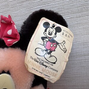 K&ouml;nnte beinhalten: Ein Minnie Mouse Pl&uuml;schtier mit einer roten Schleife mit wei&szlig;en Punkten. Das Spielzeug hat schwarze Ohren und ein rosa Gesicht. Ein Etikett mit Mickey Mouse ist angebracht, mit dem Text "MADE EXCLUSIVELY FOR THE WALT DISNEY COMPANY MINNIE MOUSE".
