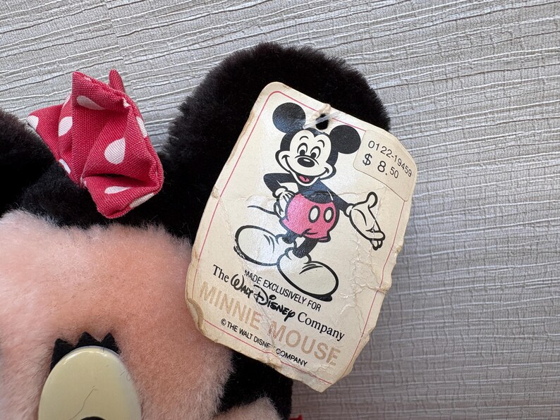 K&ouml;nnte beinhalten: Ein Minnie Mouse Pl&uuml;schtier mit einer roten Schleife mit wei&szlig;en Punkten. Das Spielzeug hat schwarze Ohren und ein rosa Gesicht. Ein Etikett mit Mickey Mouse ist angebracht, mit dem Text "MADE EXCLUSIVELY FOR THE WALT DISNEY COMPANY MINNIE MOUSE".